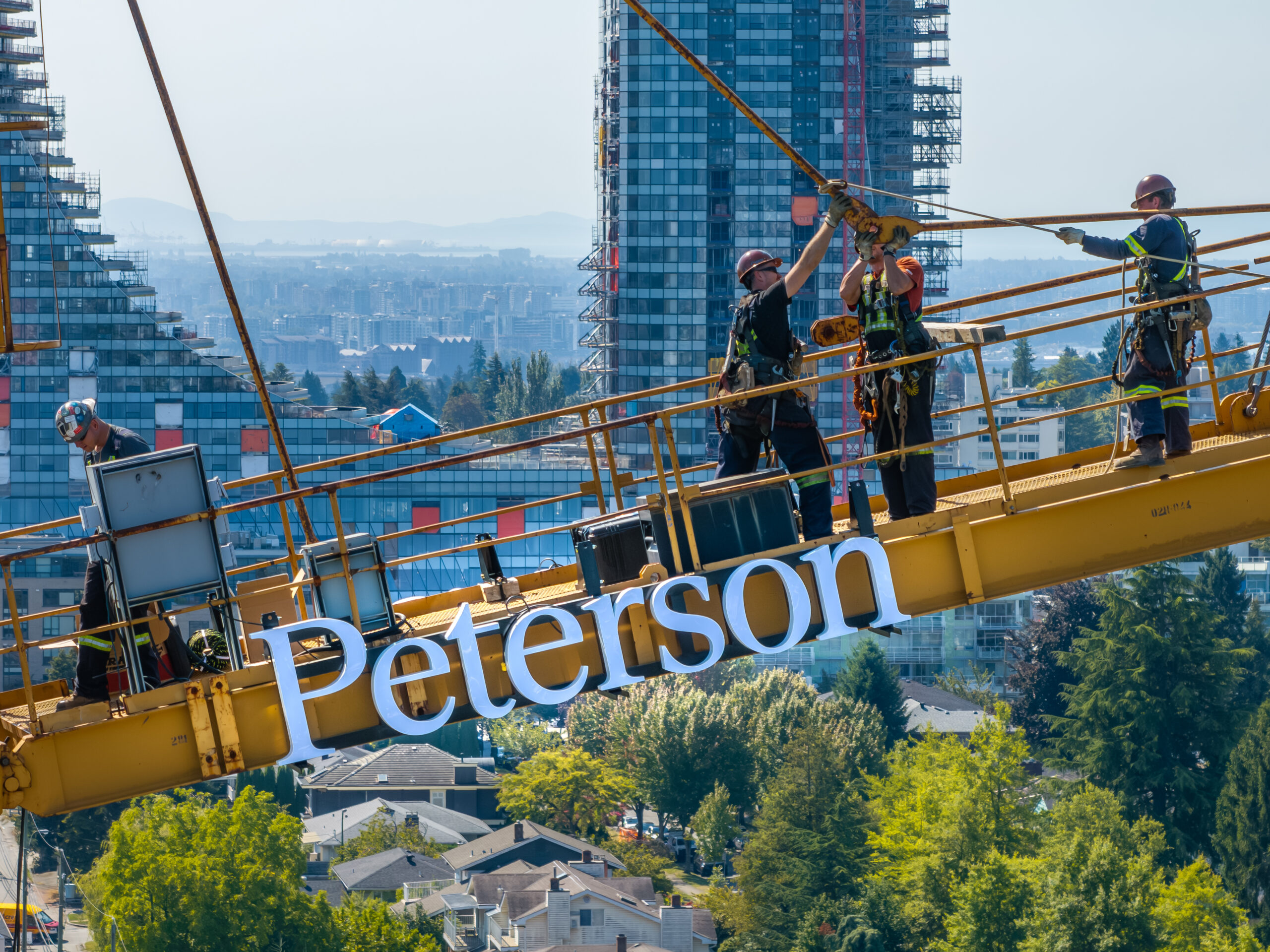 Construction workers installing a Peterson sign on a tower crane above a Vancouver construction site.
